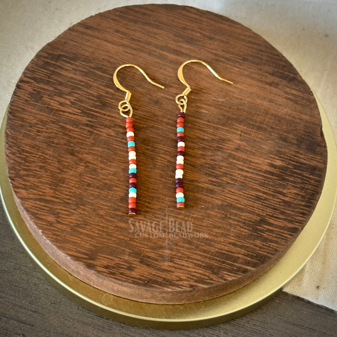A pair of handmade dangle earrings featuring vertical strands of small multicolored seed beads in shades of red, maroon, turquoise, white, and peach. The earrings hang from gold-toned fishhook ear wires and are displayed on a round, dark wooden stand with the "Savage Bead Custom Beadwork" logo faintly visible beneath.