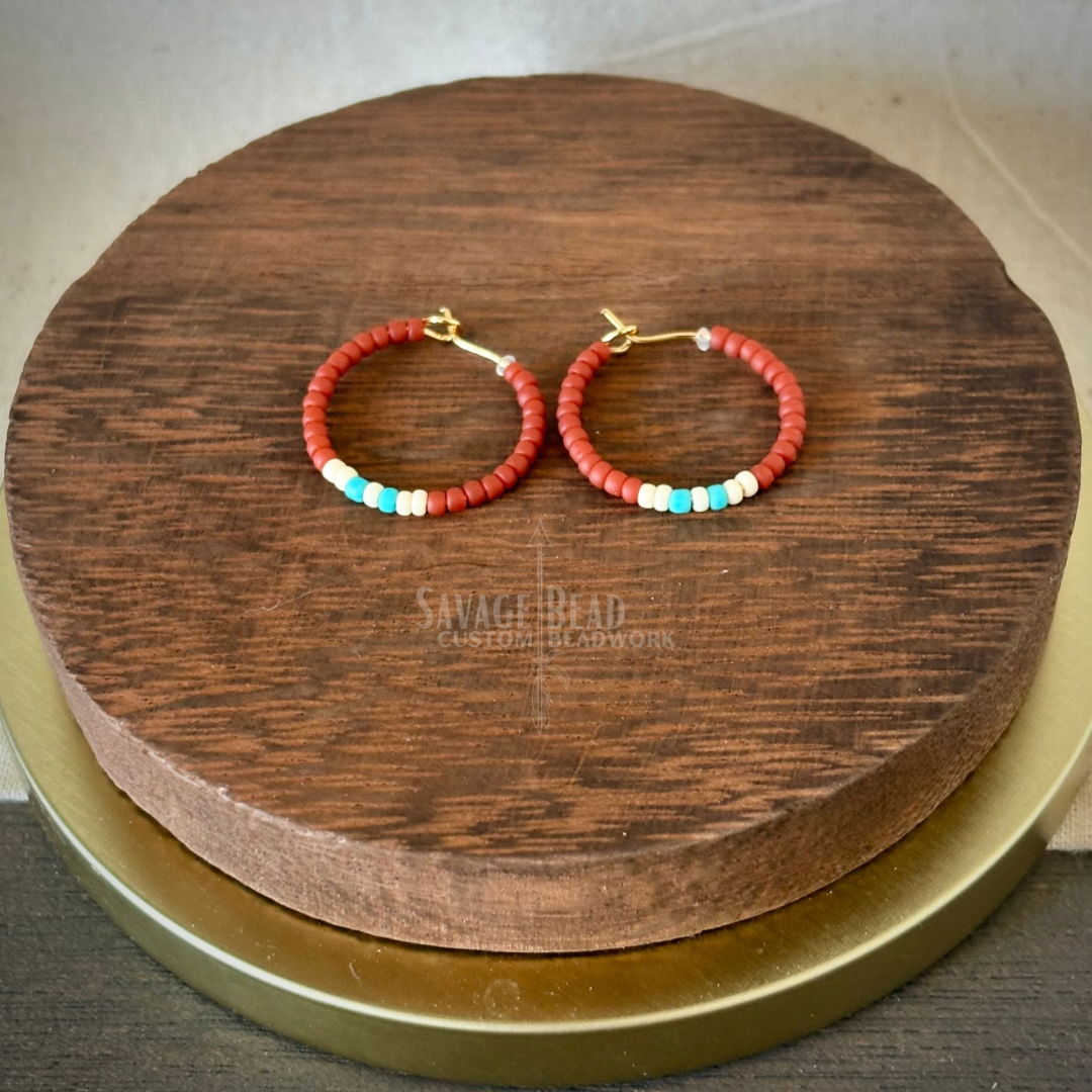 A pair of handmade hoop earrings featuring predominantly burnt orange seed beads, accented with a small section of turquoise beads. The earrings are displayed on a round dark wooden stand with the “Savage Bead Custom Beadwork” logo subtly visible underneath. The background is neutral with warm, earthy tones.