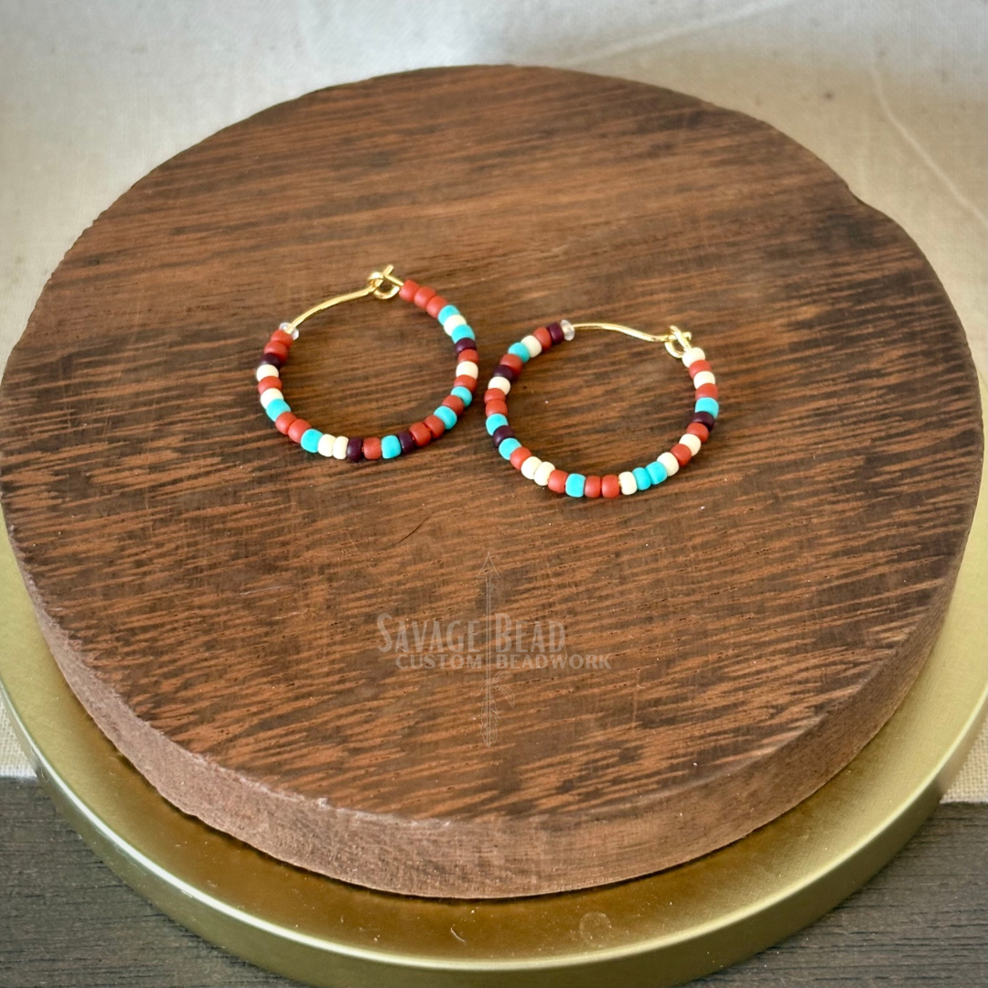 A pair of handmade hoop earrings featuring small multicolored seed beads in shades of red, turquoise, white, and burnt orange. The earrings are displayed on a round, dark wooden platform with the "Savage Bead Custom Beadwork" logo faintly visible beneath them. The background includes a soft beige and gold surface.