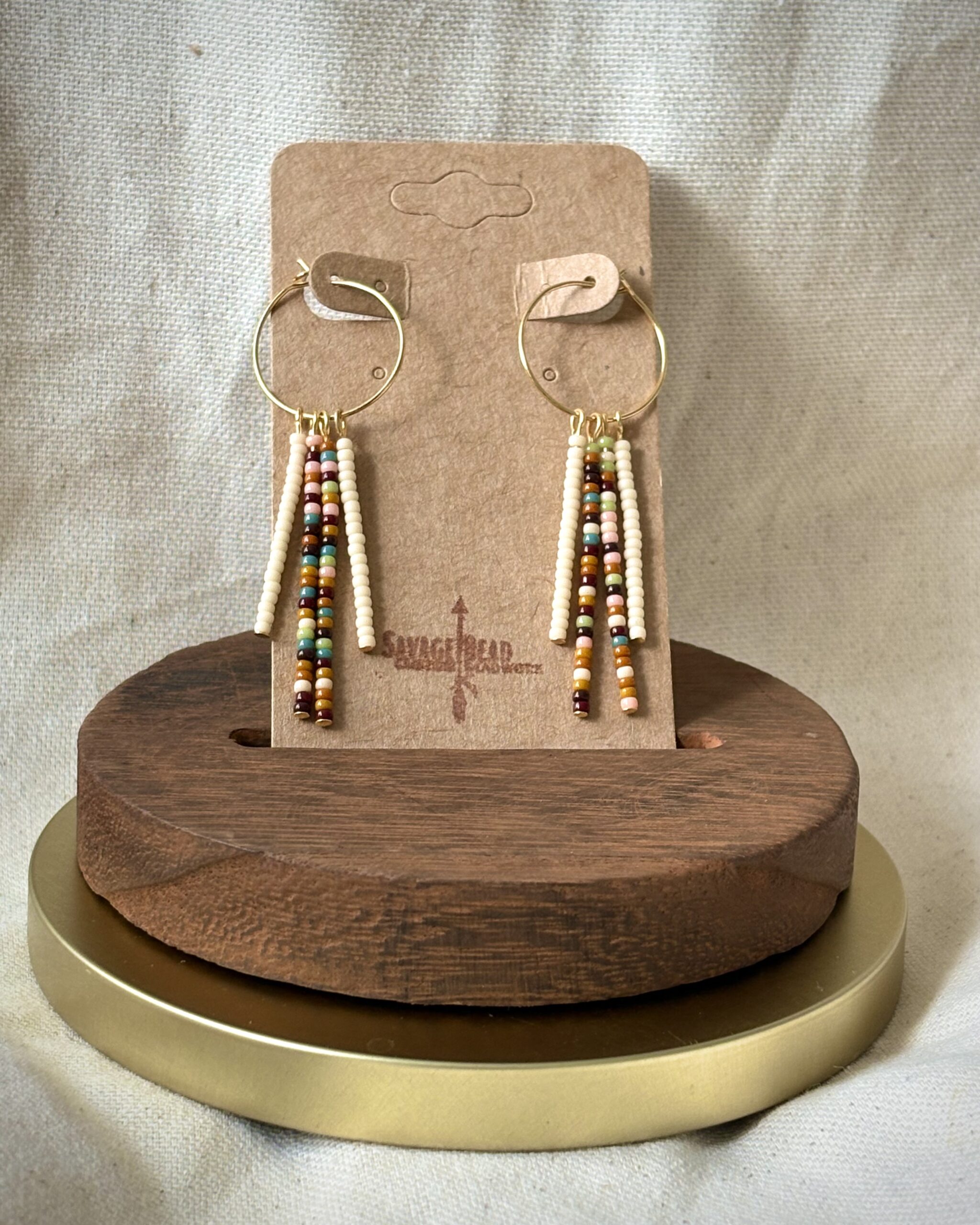 A pair of handmade boho-style dangle earrings displayed on a kraft paper card labeled "Savage Bead." The earrings feature gold hoops with 4 vertical strands of small seed beads in earthy tones including cream, rust, turquoise, mustard, pink, and brown. The card is set in a wooden stand with a gold base, against a neutral fabric background.