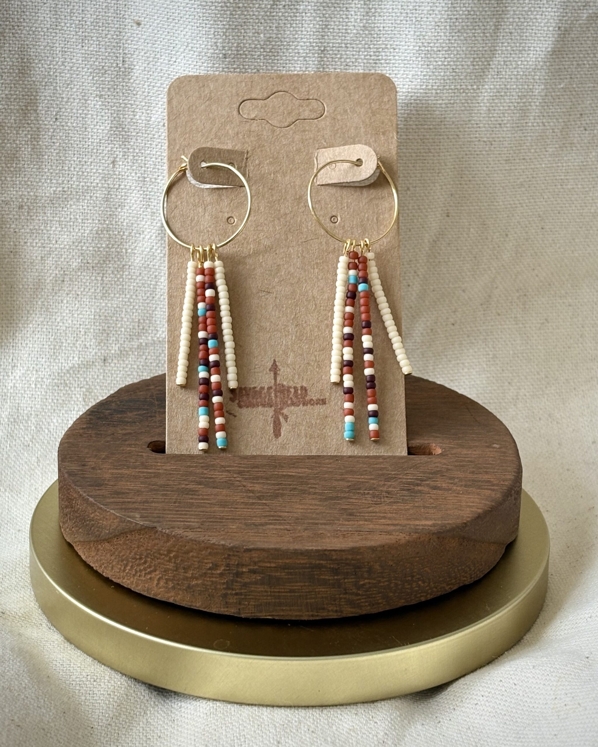 A pair of handmade savage-style dangle earrings displayed on a kraft paper card labeled "Savage Bead." The earrings feature gold hoops with three vertical strands of small seed beads in earthy tones including cream, rust, turquoise, and brown. The card is set in a wooden stand with a gold base, against a neutral fabric background.
