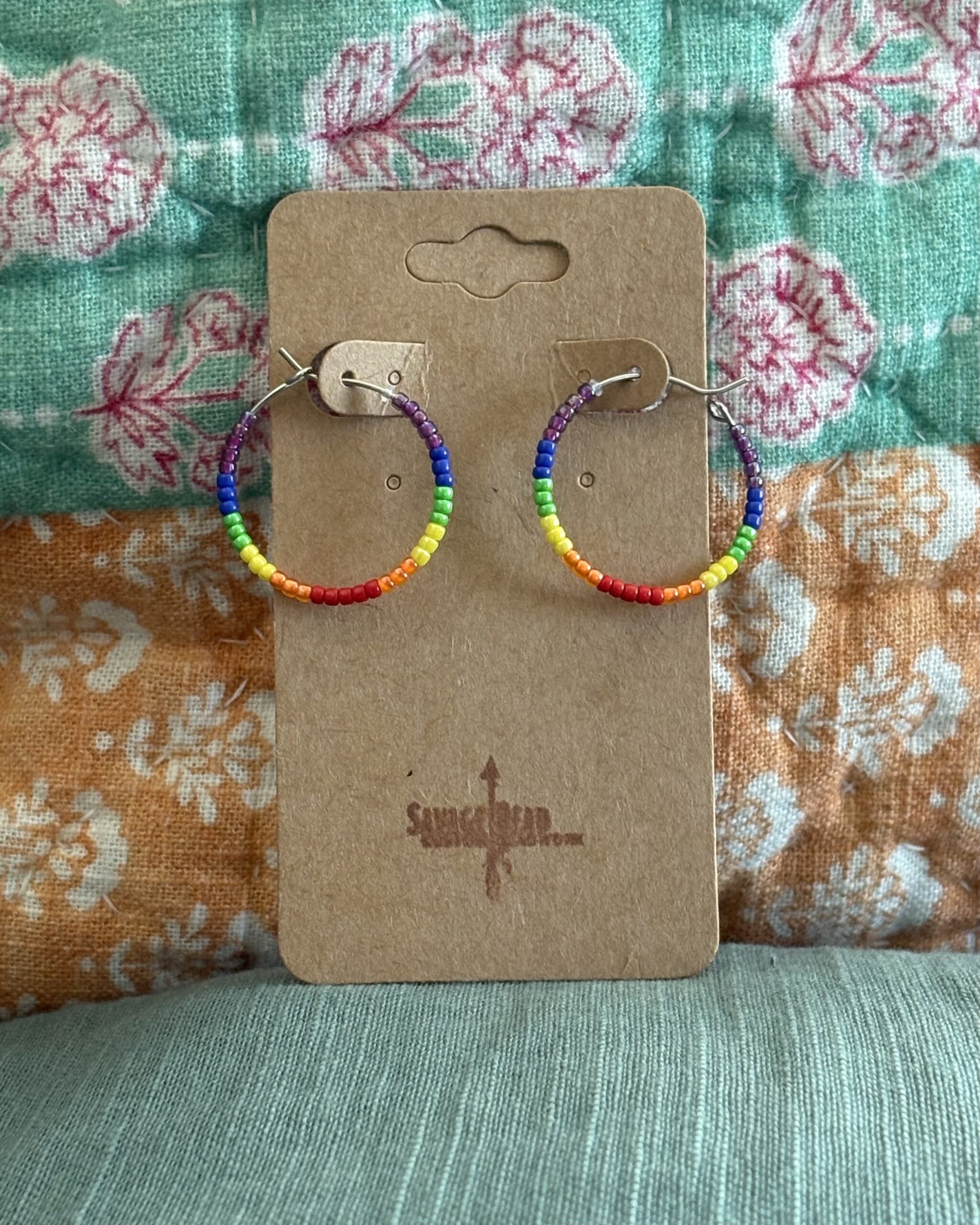 A pair of handmade hoop earrings beaded with a repeating rainbow pattern, displayed on a brown kraft card stamped with the "Savage Bead Custom Beadwork" logo. The background features a colorful, floral-patterned quilt in shades of teal, pink, orange, and white.