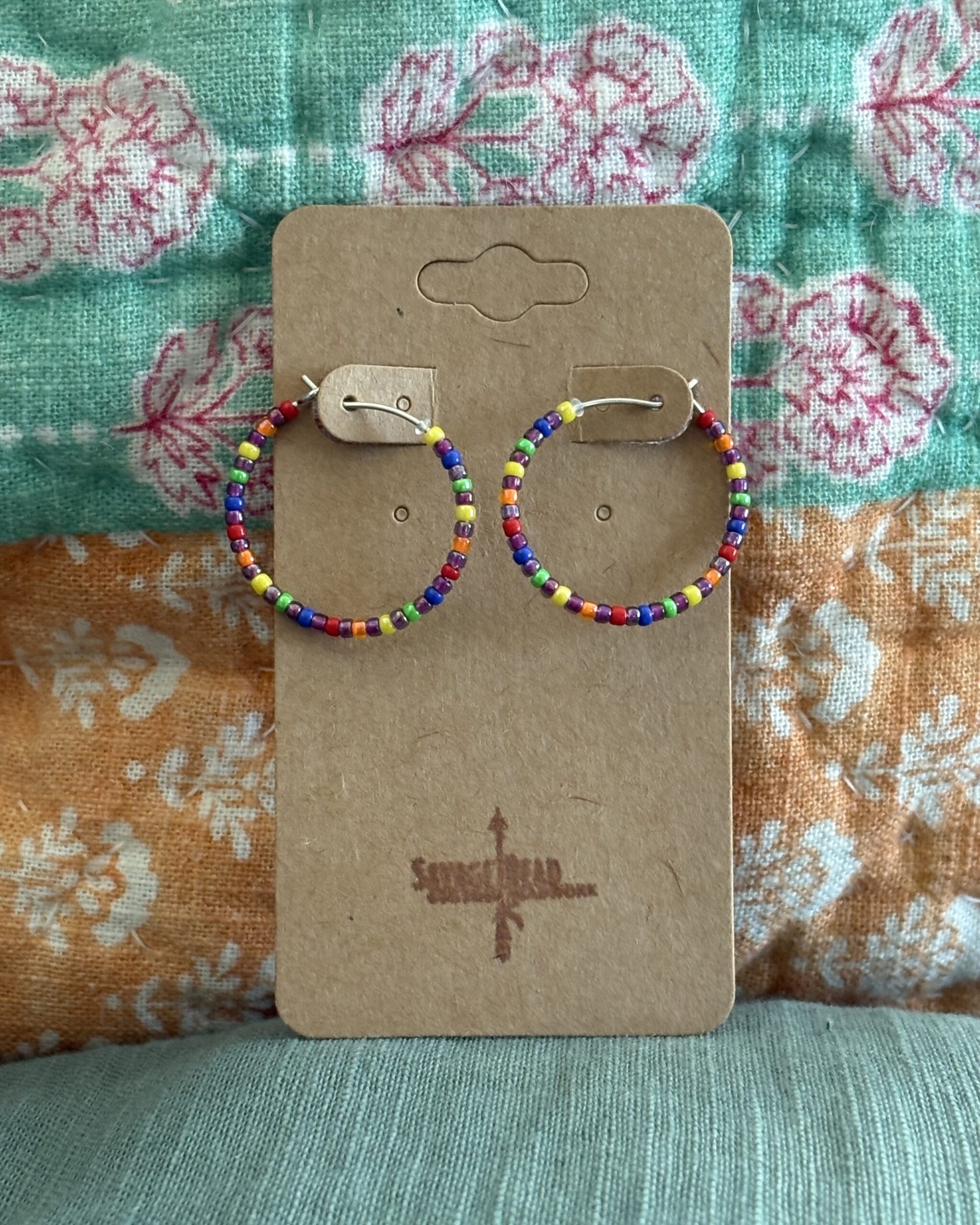 A pair of handmade hoop earrings beaded with a repeating rainbow pattern, displayed on a brown kraft card stamped with the "Savage Bead Custom Beadwork" logo. The background features a colorful, floral-patterned quilt in shades of teal, pink, orange, and white.